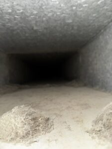 Interior view of a rectangular galvanized steel air duct heavily coated with gray dust; large tangled clumps of light fibrous debris sit in the foreground near the opening, while the far end is completely dark.
